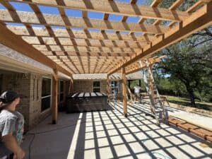 Bright image of a wooden patio cover under construction outside a brick house, with workers installing the structure.